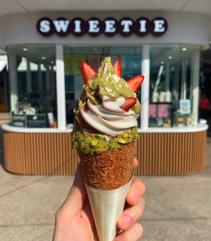 Pistachio and strawberry soft serve in a chimney cake cone, held up in front of the SWIEETIE sign